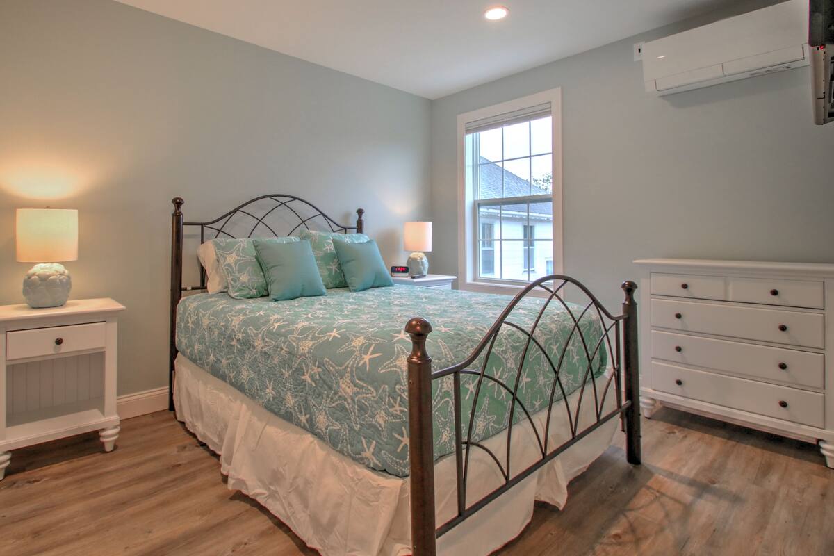 A bedroom with a bed, bedside tables, a dresser, a window, and an air conditioning unit.