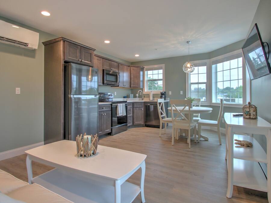 A modern kitchen with stainless steel appliances, a dining area with a white table and chairs, and large windows providing natural light.