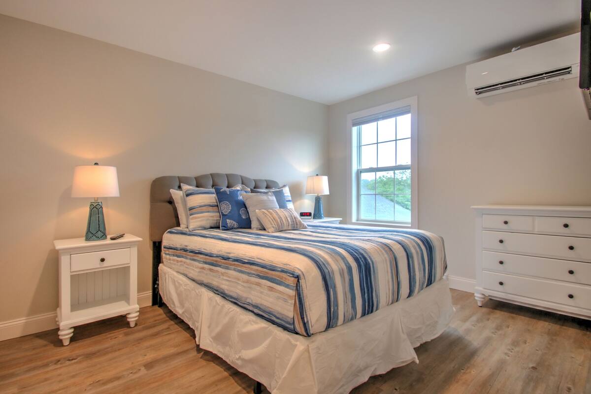 A bedroom with a large bed, two bedside tables with lamps, a dresser, and a window with a view.