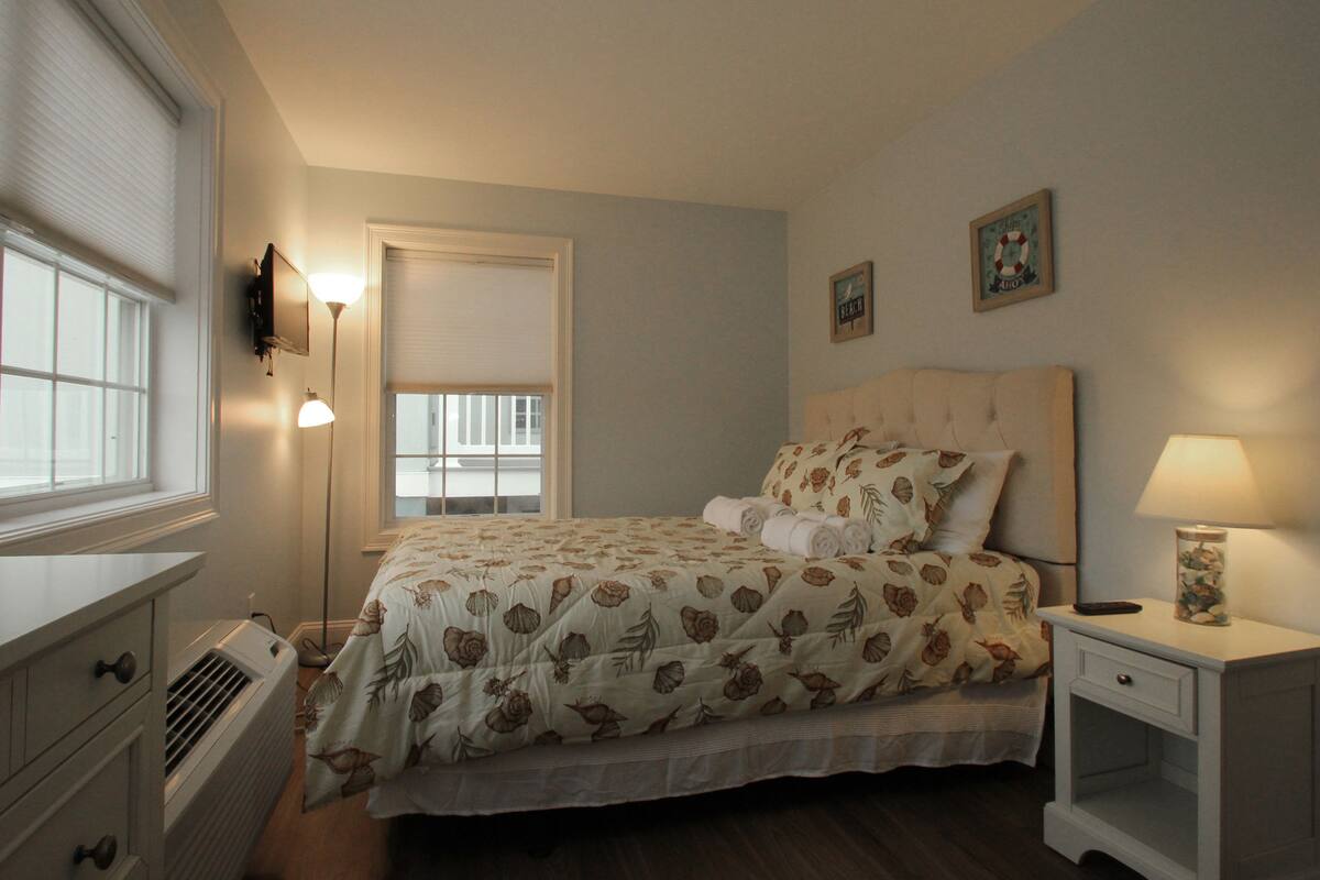 A bedroom with a bed, bedside tables, a lamp, a window, and a wall-mounted air conditioner.