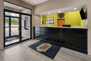 Zona de recepción con puerta de vidrio, mostrador de recepción, un reloj y una alfombra de bienvenida.
