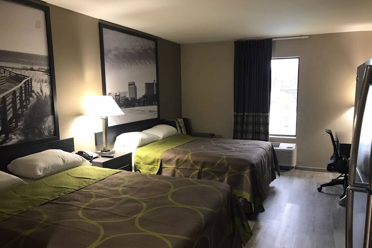 A hotel room with two beds, a desk with a lamp, a chair, a large window with curtains, and a cityscape painting on the wall.