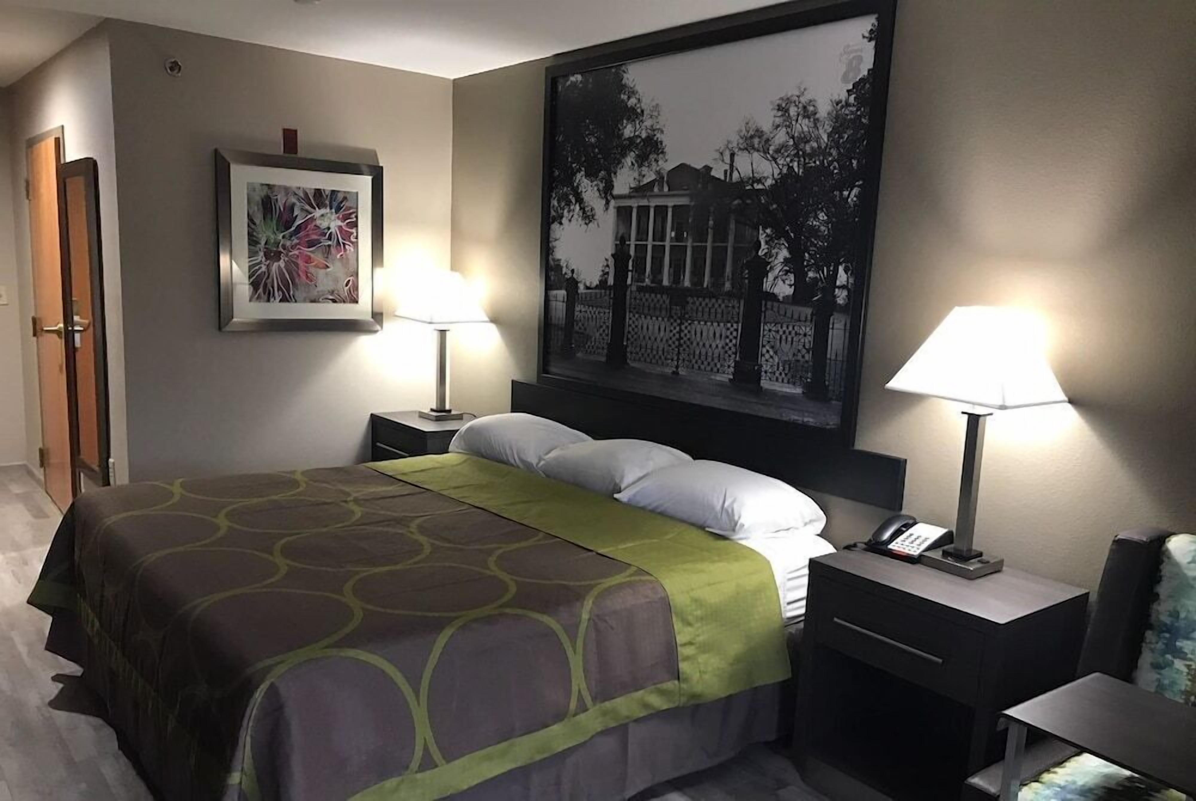 A hotel room with a bed, two bedside lamps, a nightstand with a phone, a framed picture on the wall, and a door.