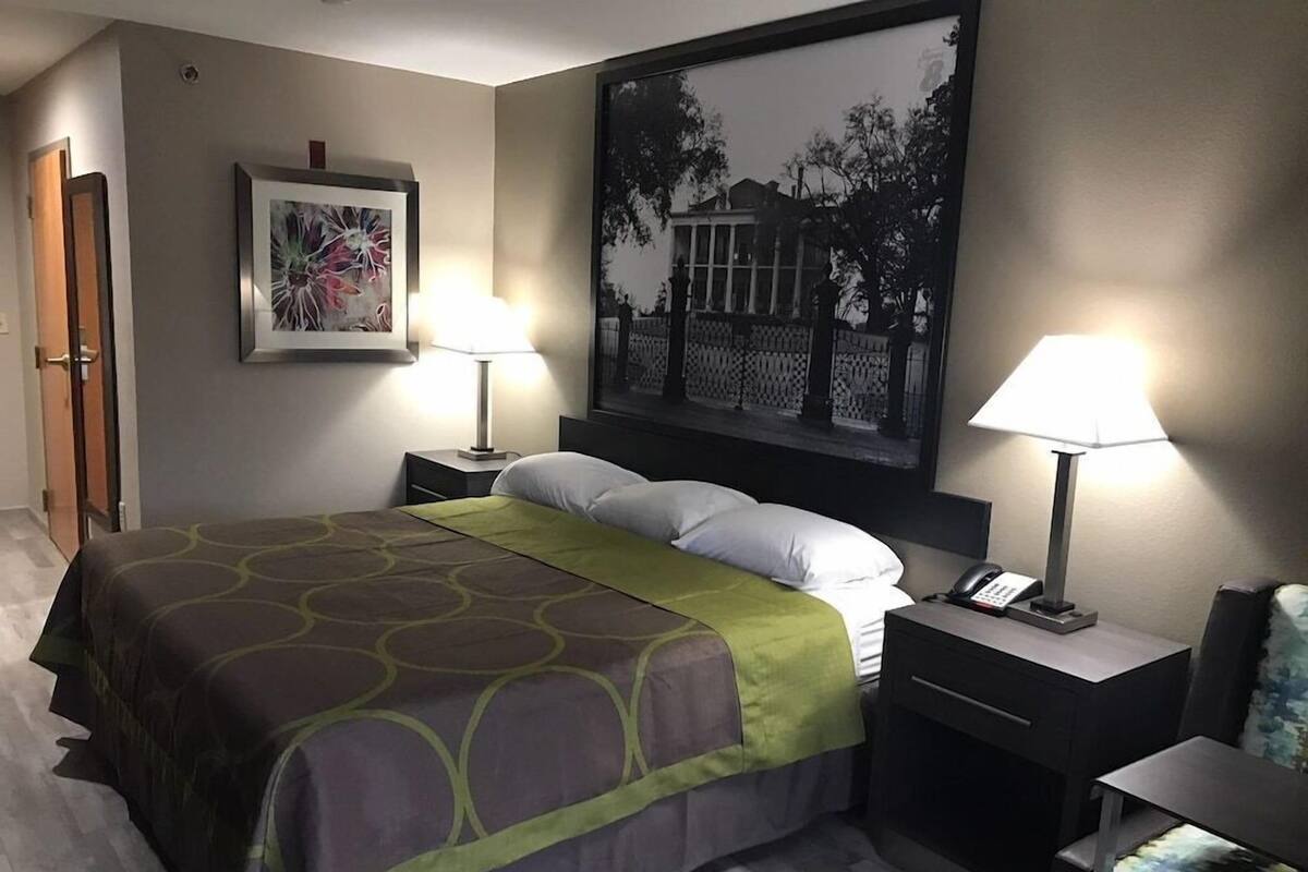 A hotel room with a bed, two bedside lamps, a nightstand with a phone, a framed picture on the wall, and a door.
