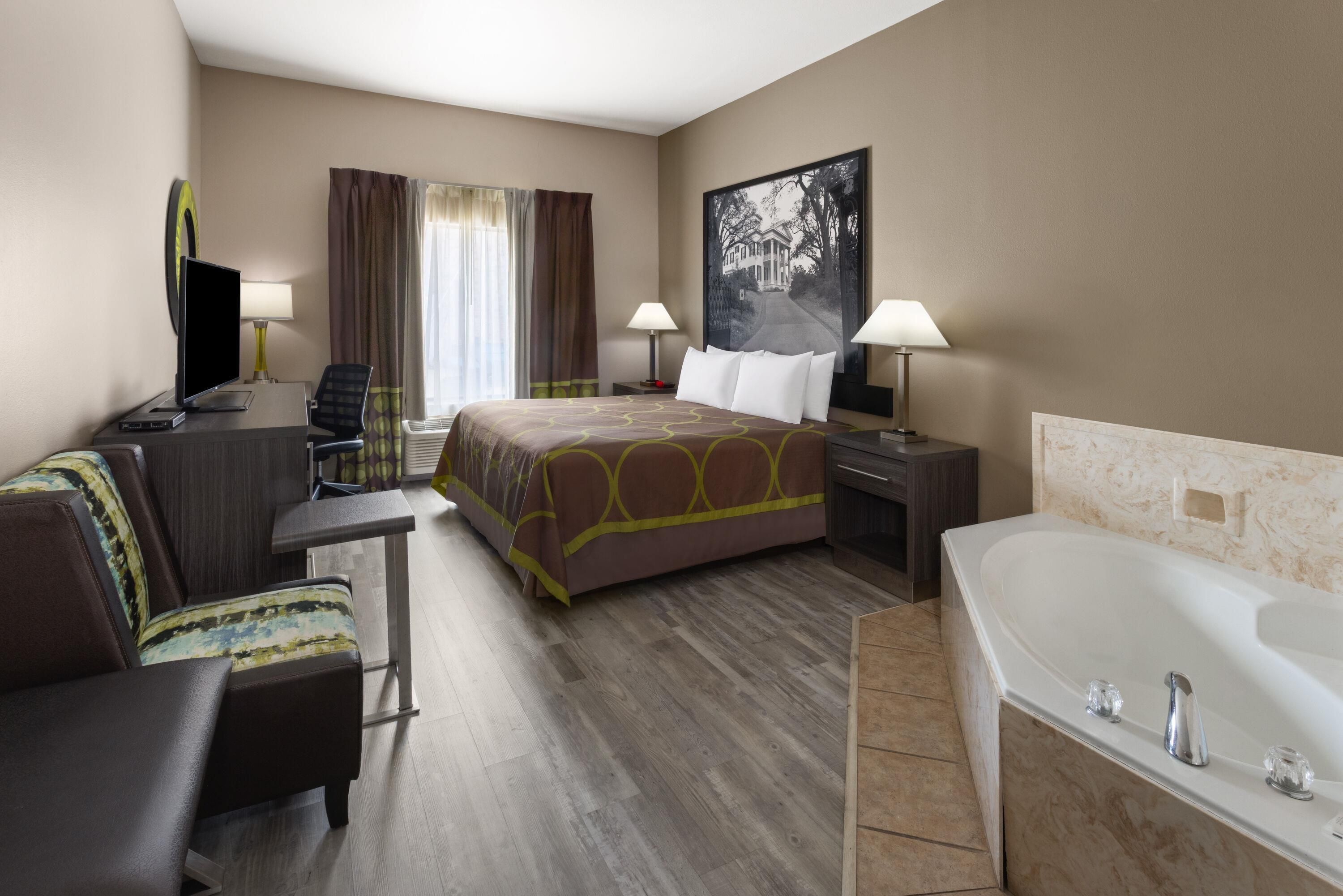 A hotel room with a bed, a desk, a chair, a TV, a lamp, a painting, and a bathtub.