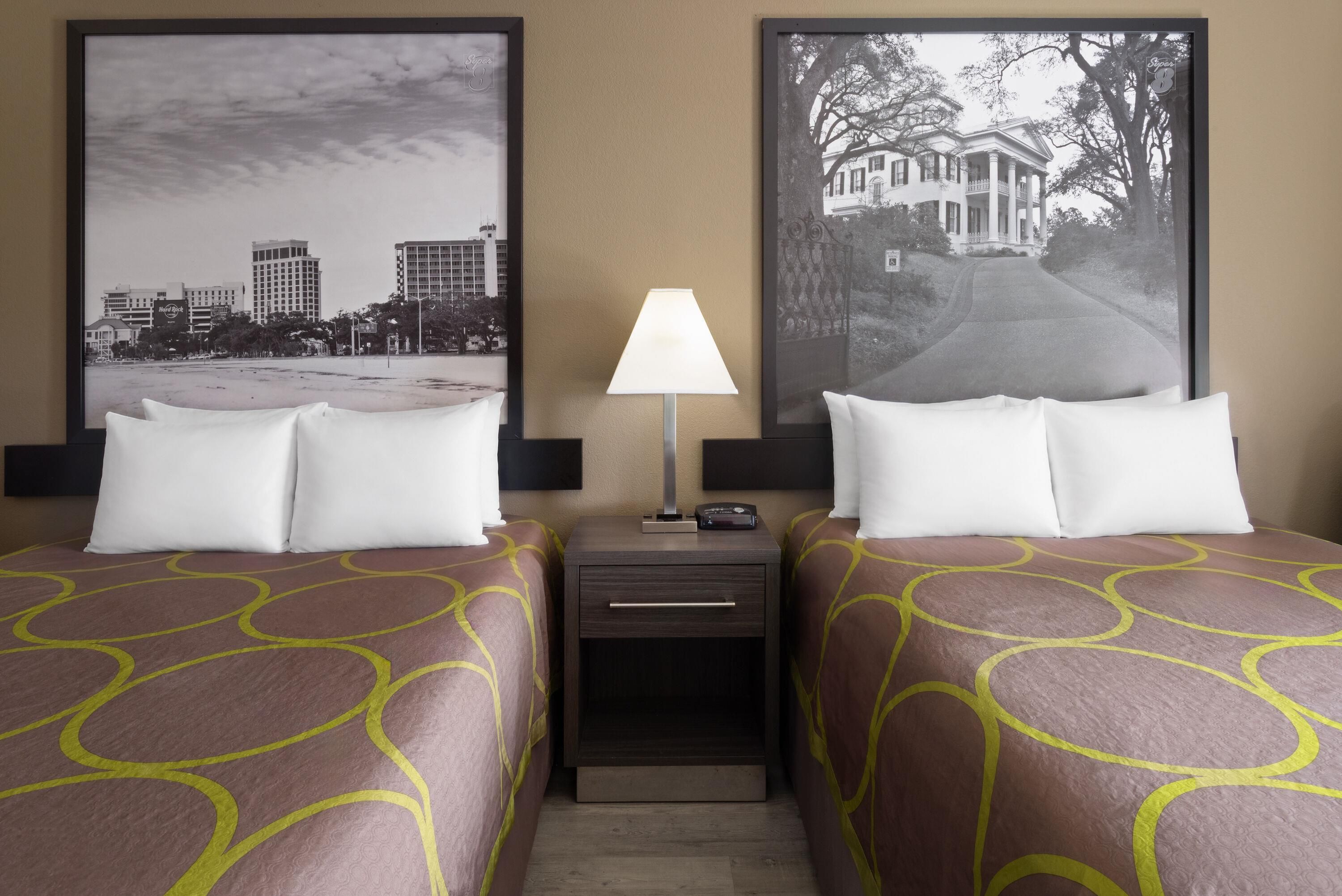 Habitación de hotel con dos camas, una mesita de noche, una lámpara y dos cuadros en la pared.