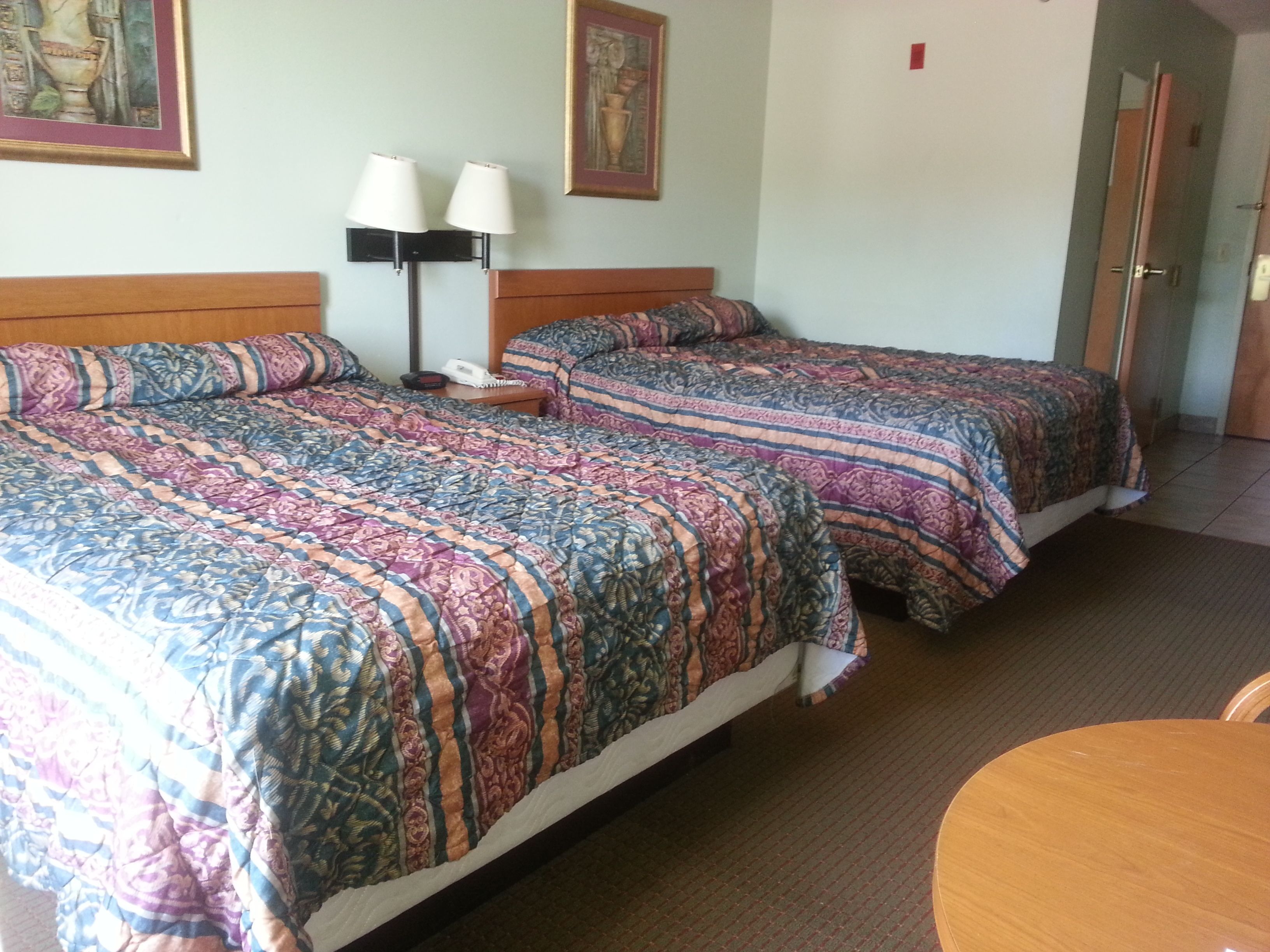 A hotel room with two beds, each with patterned bedding, a bedside table with lamps, and a painting on the wall.