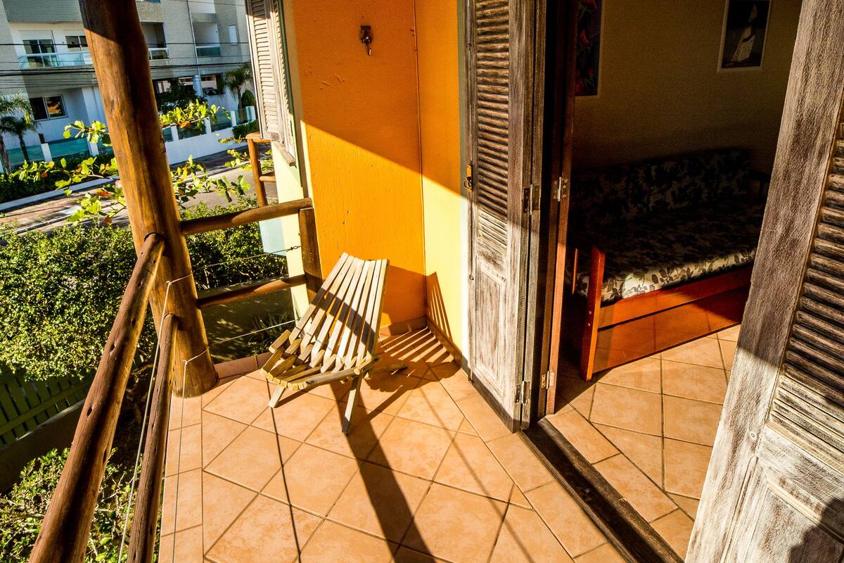 Un balcón con una silla de madera, una cama y vistas a un edificio y follaje.