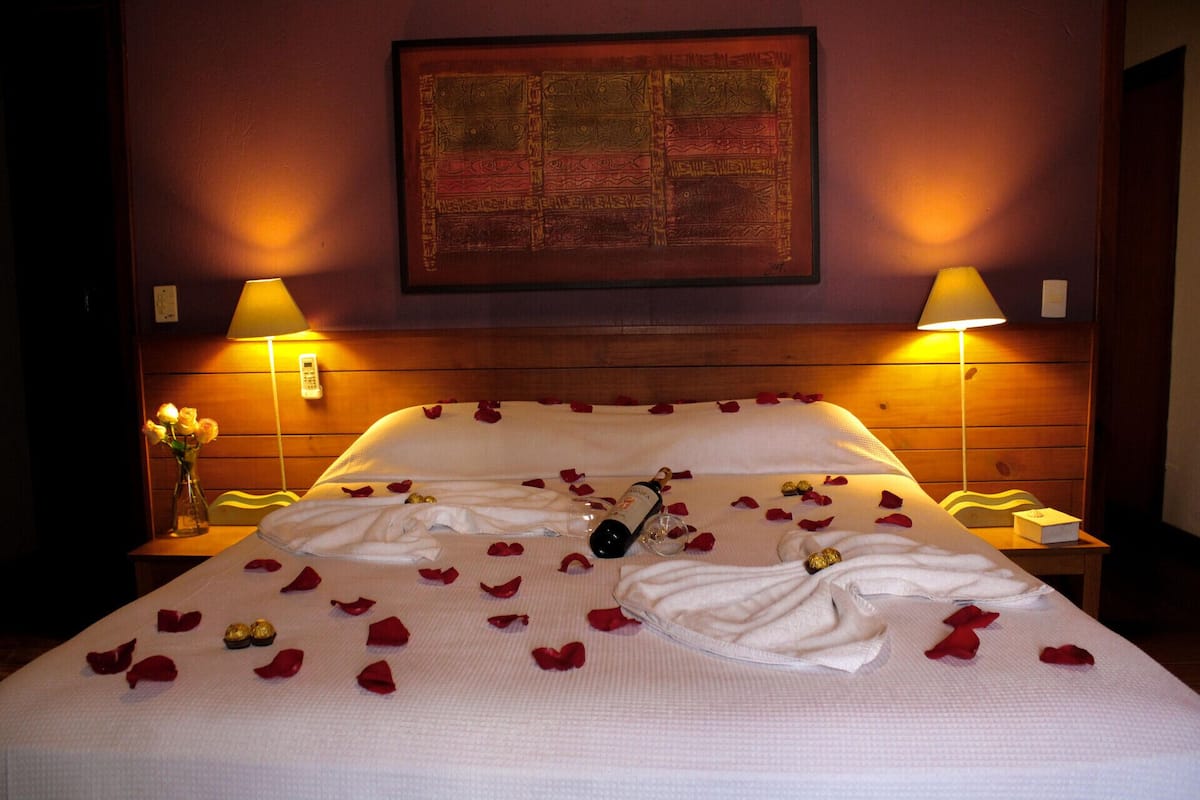 Habitación de hotel con una cama, pétalos de rosa, una botella y una obra de arte enmarcada en la pared.