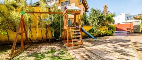 A wooden deck with a swing set and a slide, surrounded by lush greenery and a yellow wall.