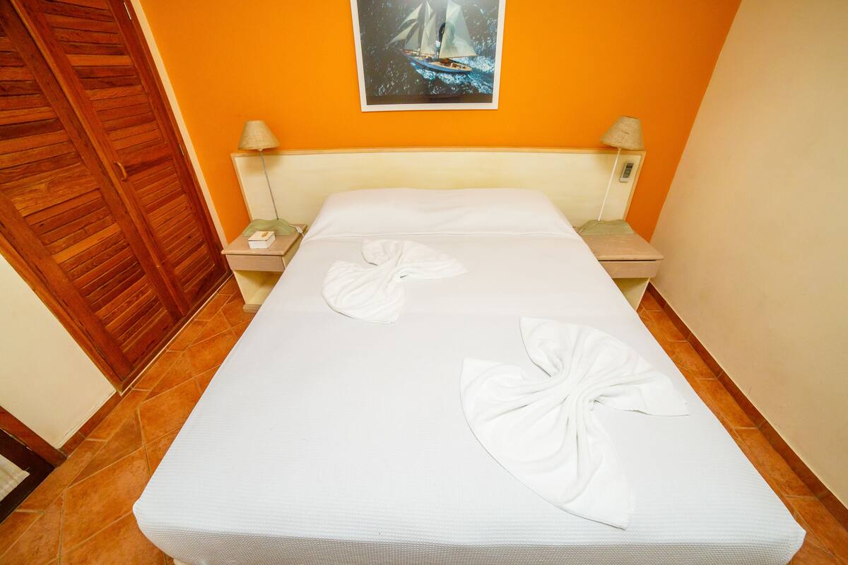 Una cama bien hecha con sábanas blancas y dos toallas, un armario de madera y un cuadro de un velero en la pared.