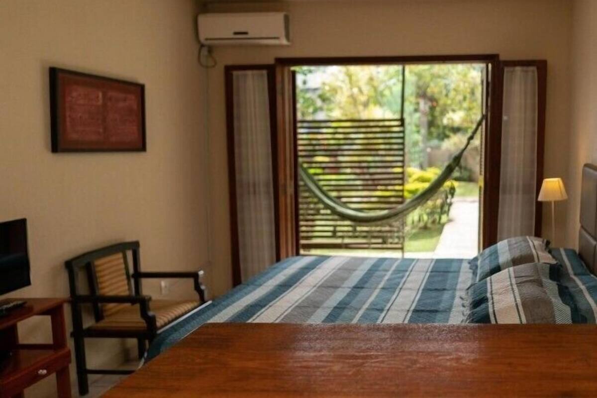 Un dormitorio con cama, una silla, un televisor, una ventana con vista a follaje, y una hamaca afuera.