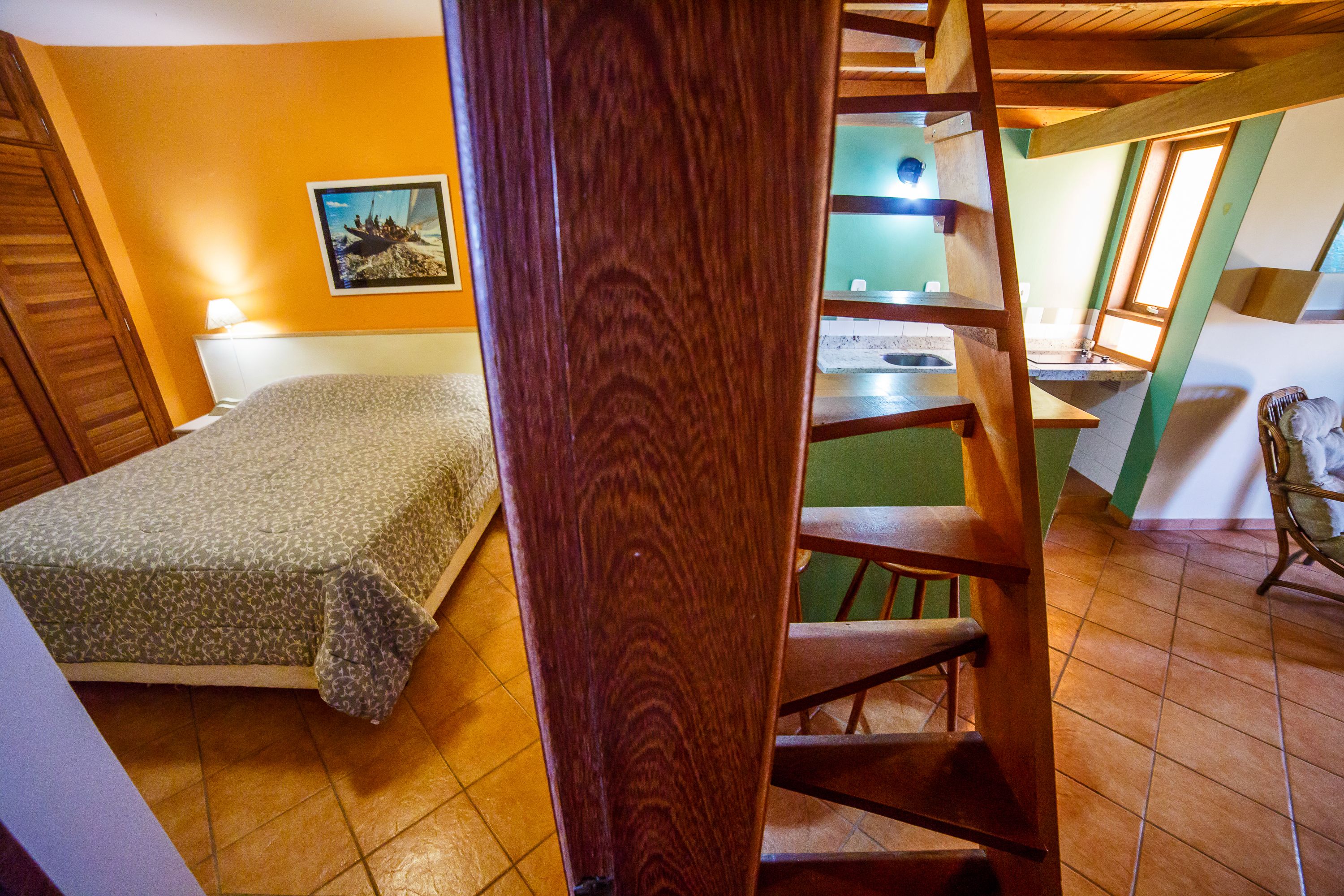 A bedroom with a bed, a wooden chair, a staircase, and a bathroom with a sink and mirror.
