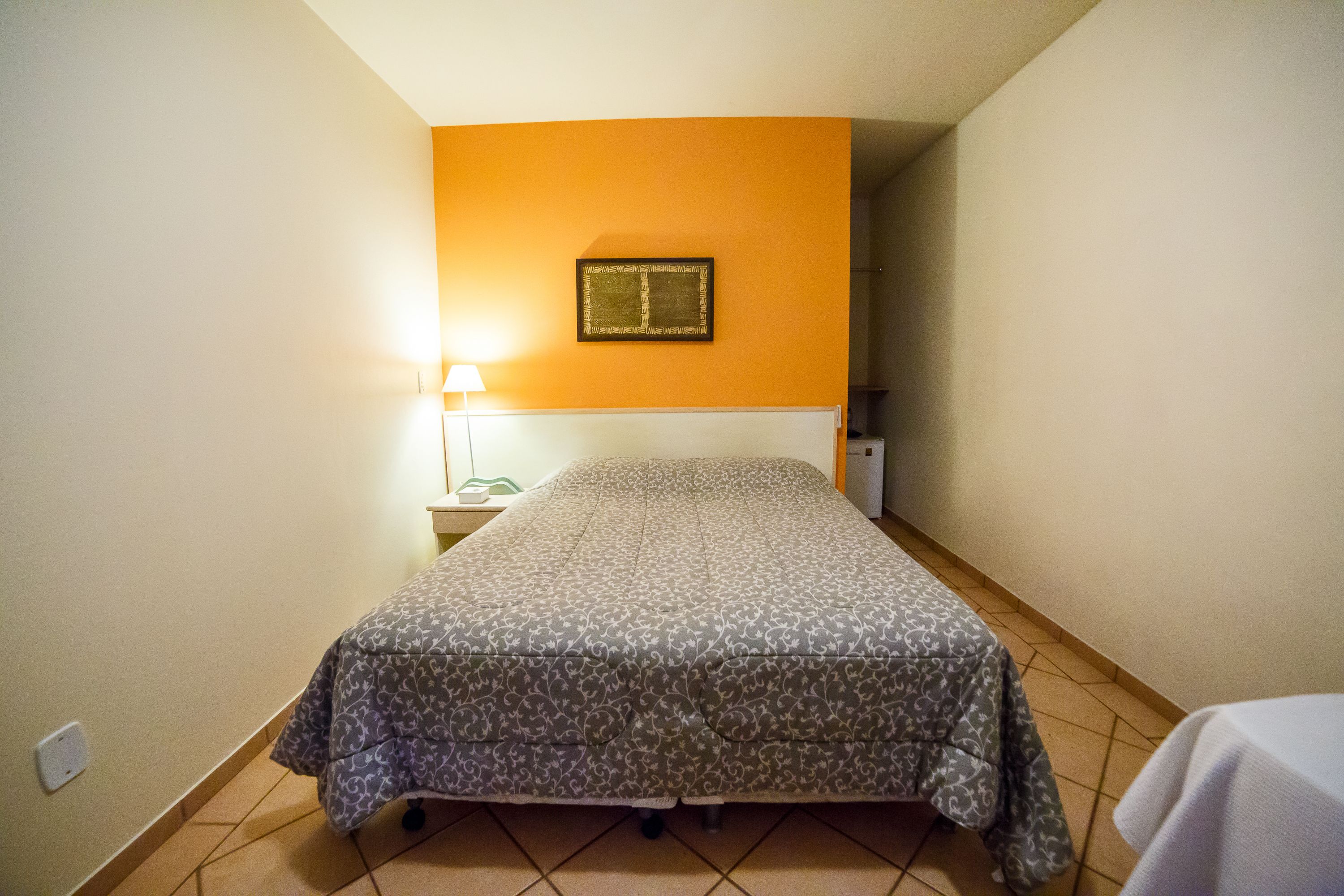 A small, narrow room with a bed, a nightstand, a lamp, and a framed picture on the wall.