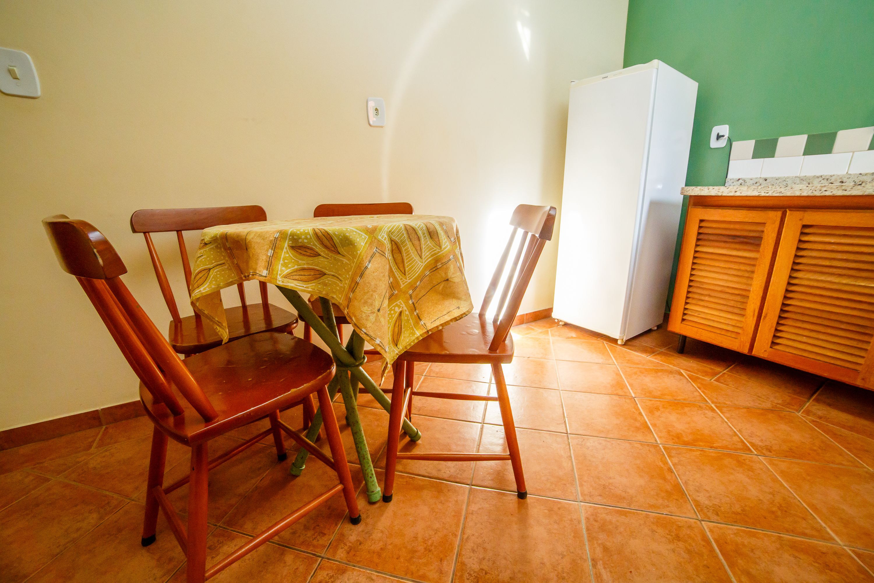 A dining area with a table covered by a yellow tablecloth, wooden chairs, and a white refrigerator in the background.