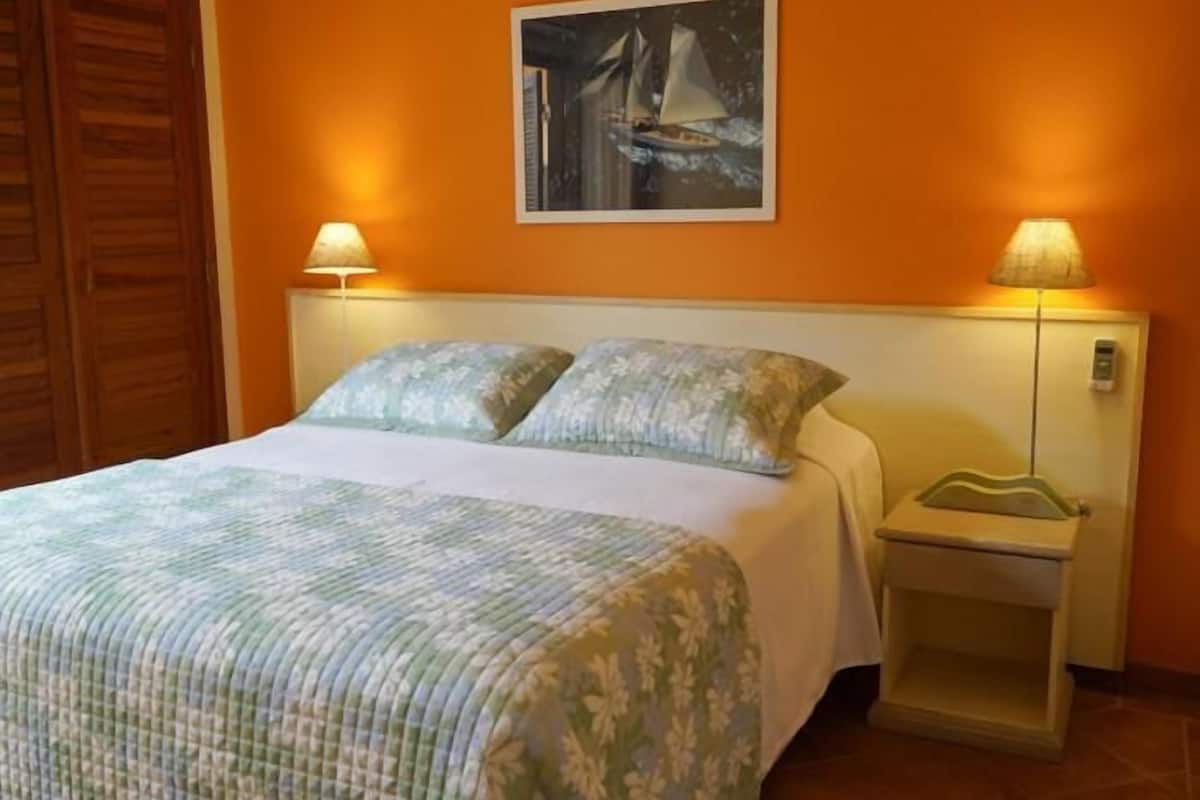 Una cama con una colcha estampada con motivos florales, dos lámparas de noche, una mesita de noche con lámpara y un cuadro de un velero en la pared.