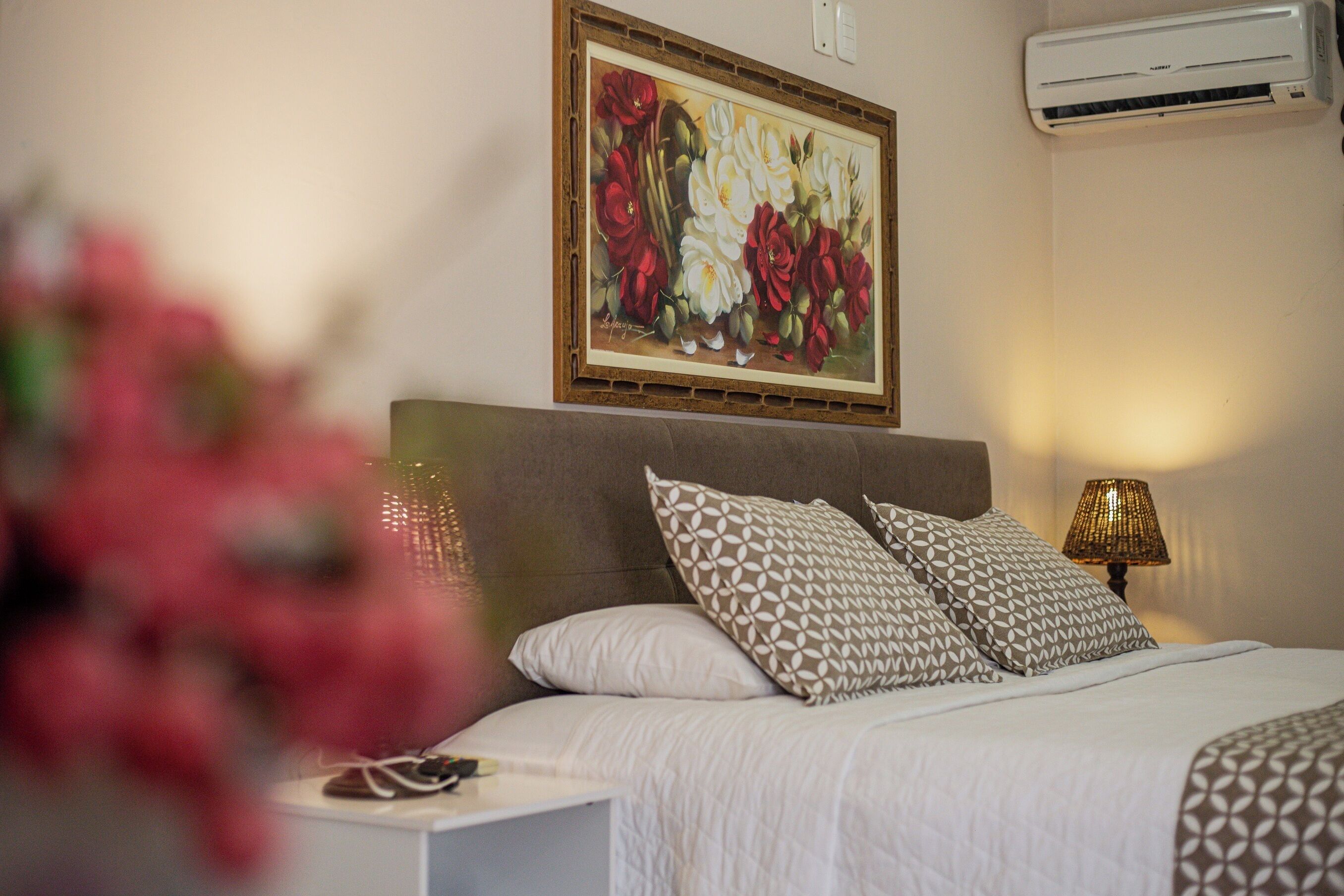 A neatly made bed with patterned pillows, a framed floral artwork above it, and a bedside lamp.