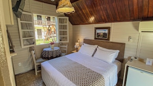A cozy bedroom with a bed, a small table with a checkered tablecloth, a chair, a nightstand with a lamp, and a window with a view of outdoor furniture. - Pousada Caliandra da Serra (Canela)