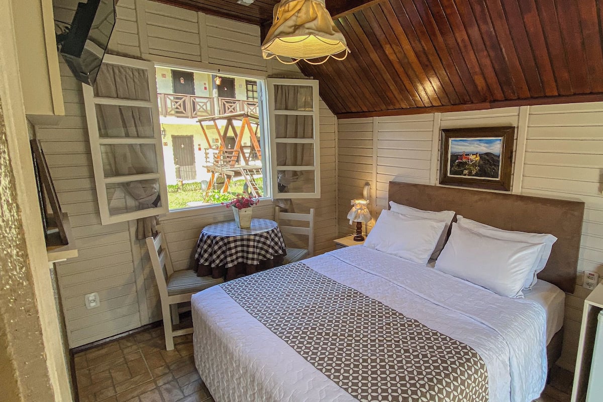Ein gemütliches Schlafzimmer mit einem Bett, einem kleinen Tisch mit karierter Tischdecke, einem Sessel, einem Nachttisch mit Lampe und einem Fenster mit Blick auf Gartenmöbel.