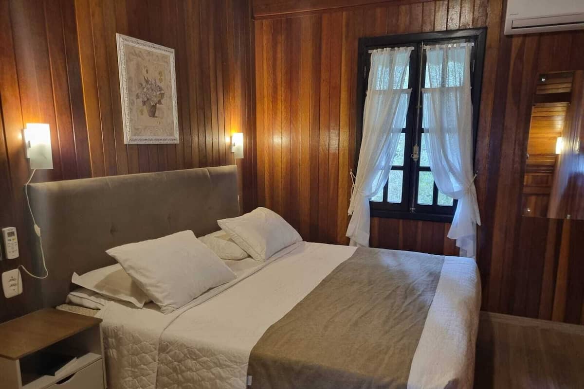 Ein ordentlich bezogenes Bett mit gesteppter Bettdecke und Kissen, ein Nachttisch mit Lampe sowie ein Fenster mit durchsichtigen Vorhängen.