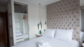 A neatly made bed with white linens and towels, a tufted headboard, a white wardrobe with a mirror, and a small bedside table with a lamp. - Residencial Florence (Gramado)