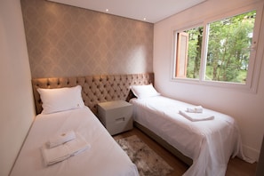 A bedroom with two beds, a nightstand, and a window with a view of trees. - Residencial Florence (Gramado)