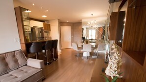 A modern living room with a sofa, dining area with chairs, a kitchen with a refrigerator, and a wooden reception desk. - Residencial Florence (Gramado)