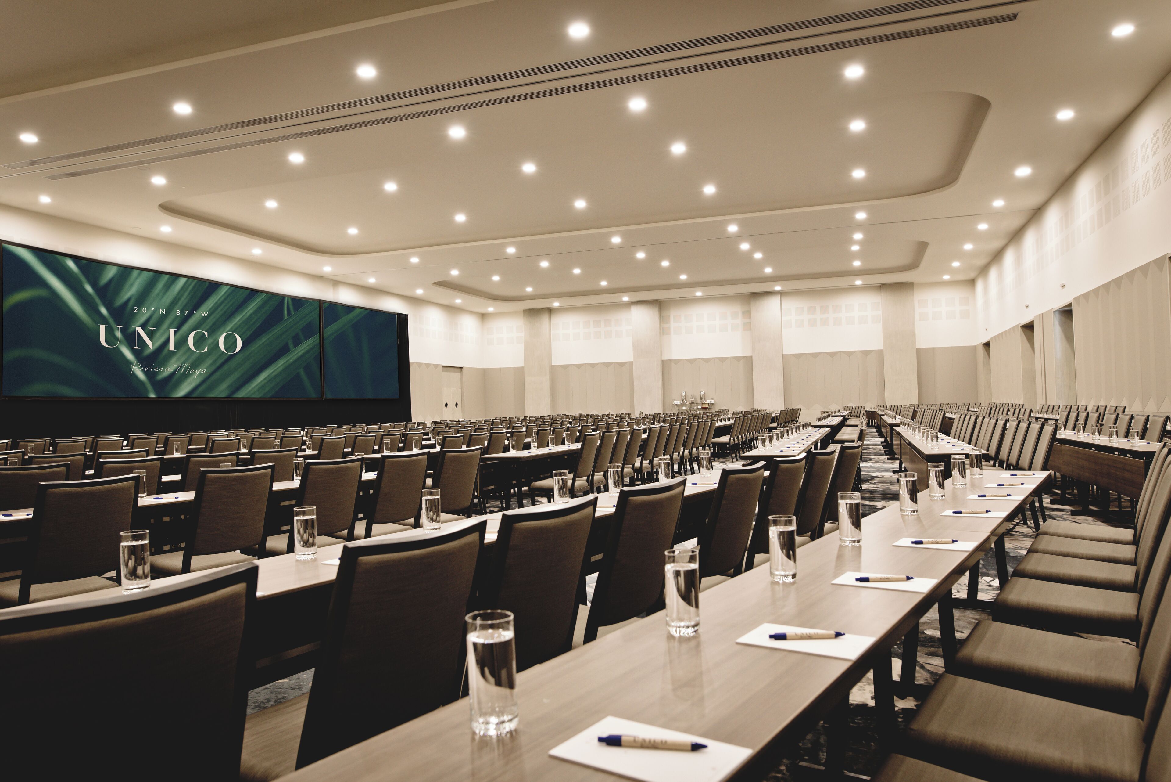 A conference room with rows of tables and chairs, a large screen displaying 'UNICO', and a modern ceiling design.