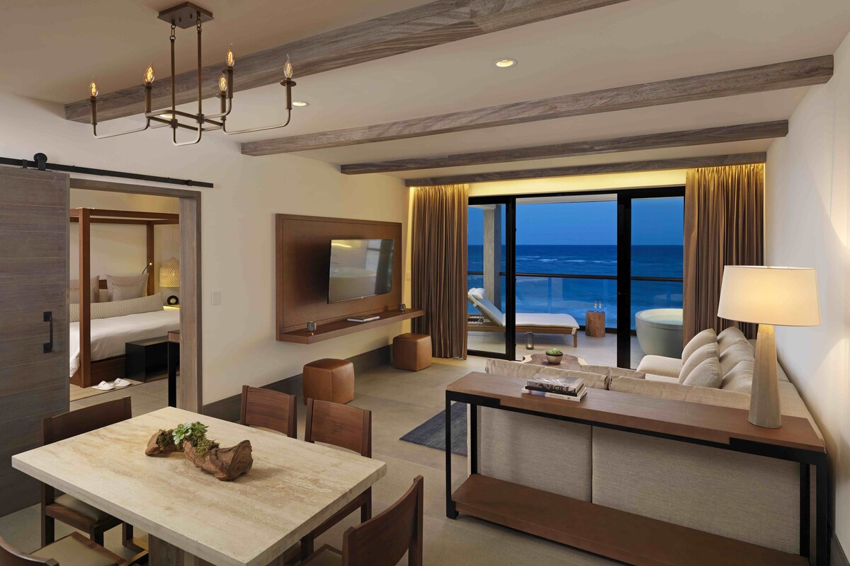 A hotel room with a balcony, a dining area, a TV, and a seating area with a view of the ocean.