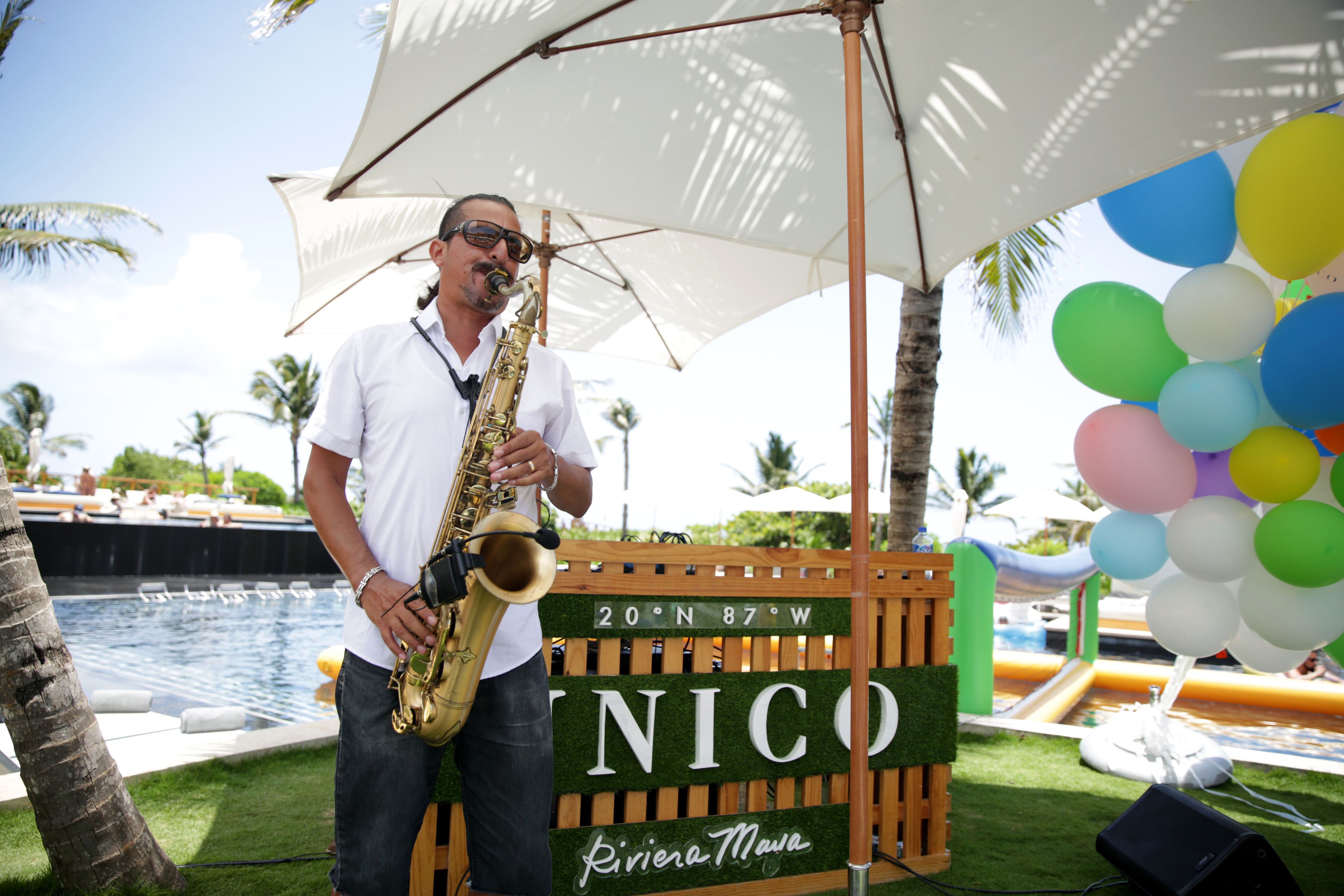 A man playing a saxophone under a white umbrella by a poolside with a sign reading "20ºN 87ºW Riviera Maya".