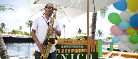 A man playing a saxophone under a white umbrella by a poolside with a sign reading "20ºN 87ºW Riviera Maya".