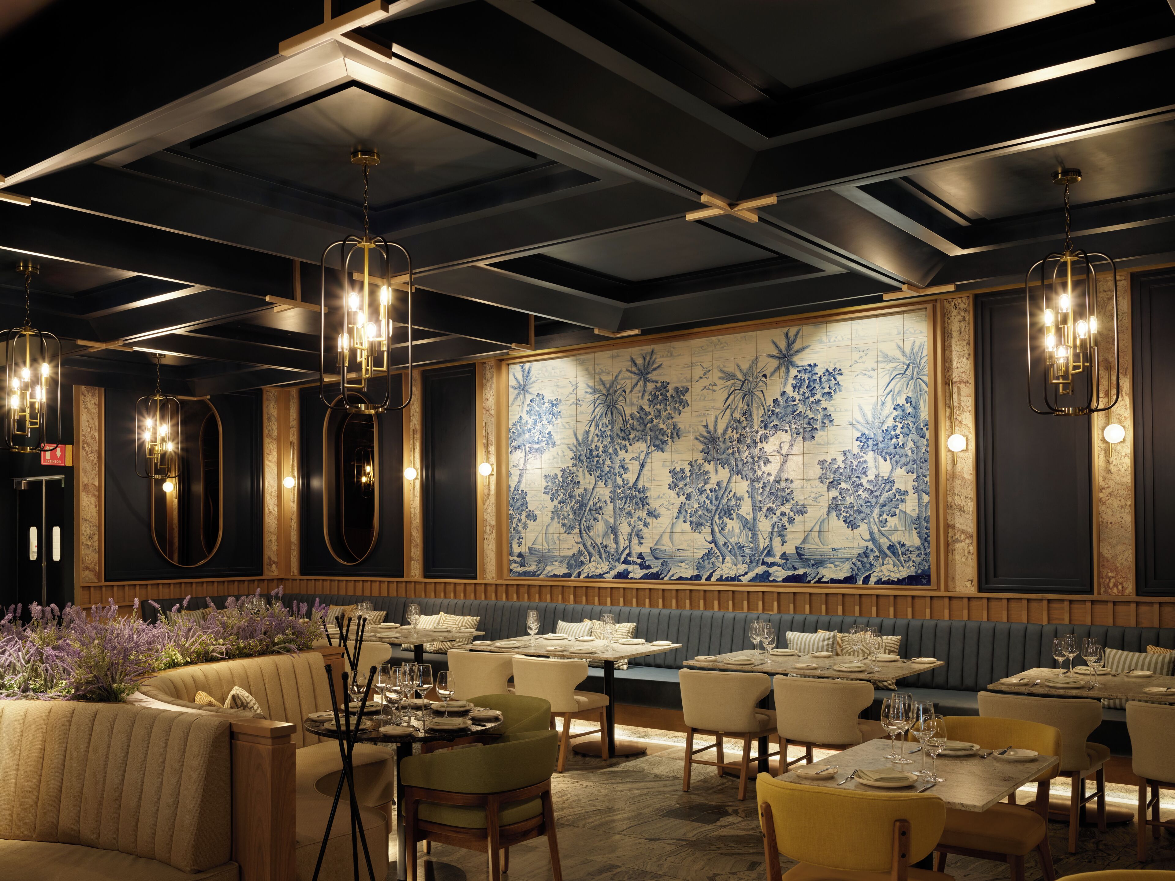 Ein Restaurant mit hohen Decken, Pendelleuchten und einer großen Wanddekoration. Die Tische sind mit weißen Tischdecken und Kerzen gedeckt.