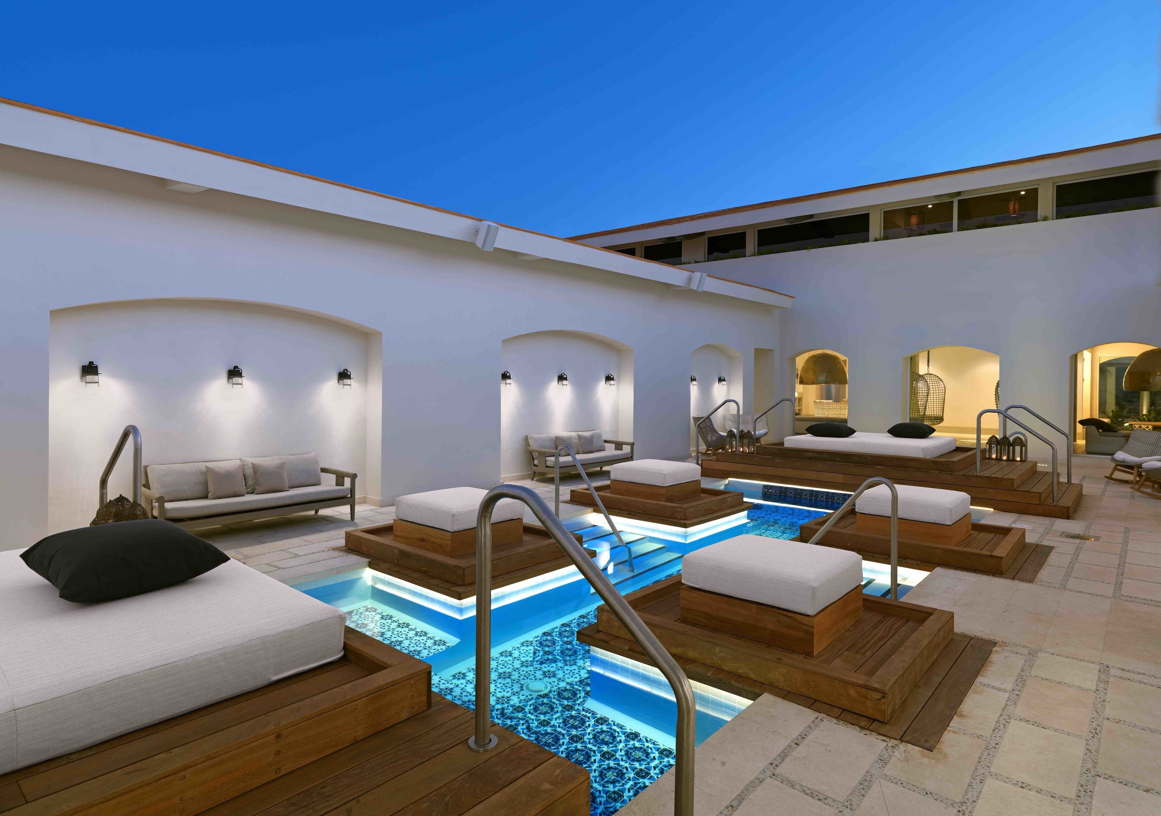 Zona de piscina con tumbonas de madera y una piscina pequeña, rodeada de paredes blancas y espacios de descanso.