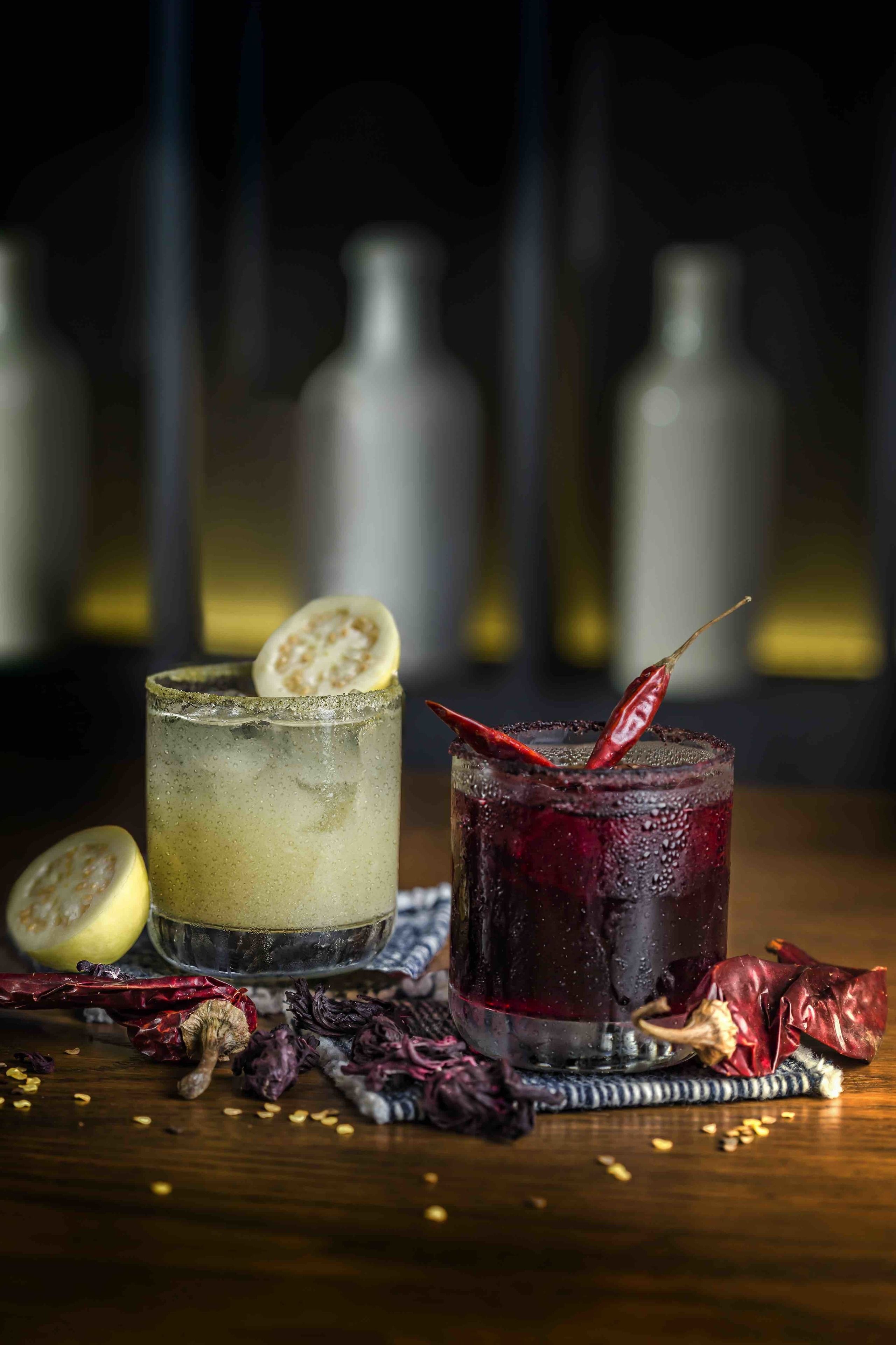 Two glasses of beverages on a wooden surface with dried chili peppers and a lemon slice.