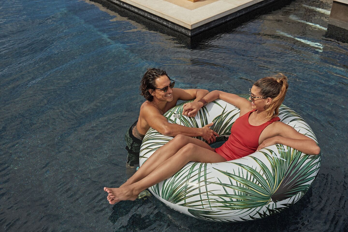 Dos personas sentadas en un flotador inflable con diseño de hojas en una piscina.