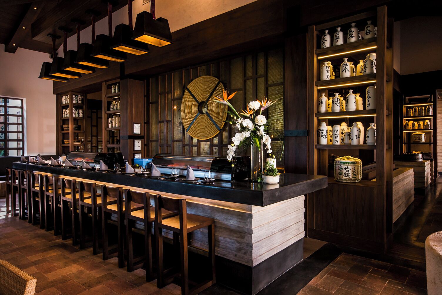 A bar area with a long counter, multiple stools, and shelves with bottles and jars.