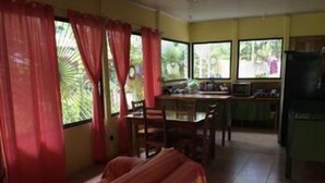 A room with a bed, a dining table with chairs, a kitchen area with a refrigerator, and large windows with curtains. - La Casita del Sol (Cahuita)