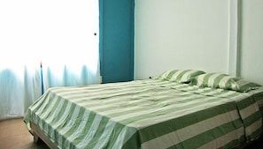 Un lit simple, recouvert d’une couette rayée verte et blanche, avec des coussins assortis, dans une pièce où l’on trouve un rideau bleu et un mur blanc.