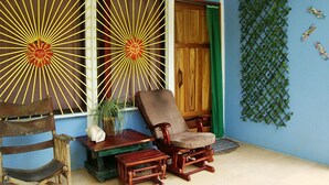 A room with a wooden rocking chair, a small table, and a bench. There is a window with a sunburst design and a door with a wooden panel. - La Casita del Sol (Cahuita)
