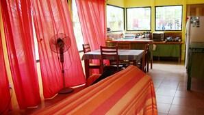 A room with a bed, a fan, a table, chairs, a kitchen area, and a window with curtains. - La Casita del Sol (Cahuita)