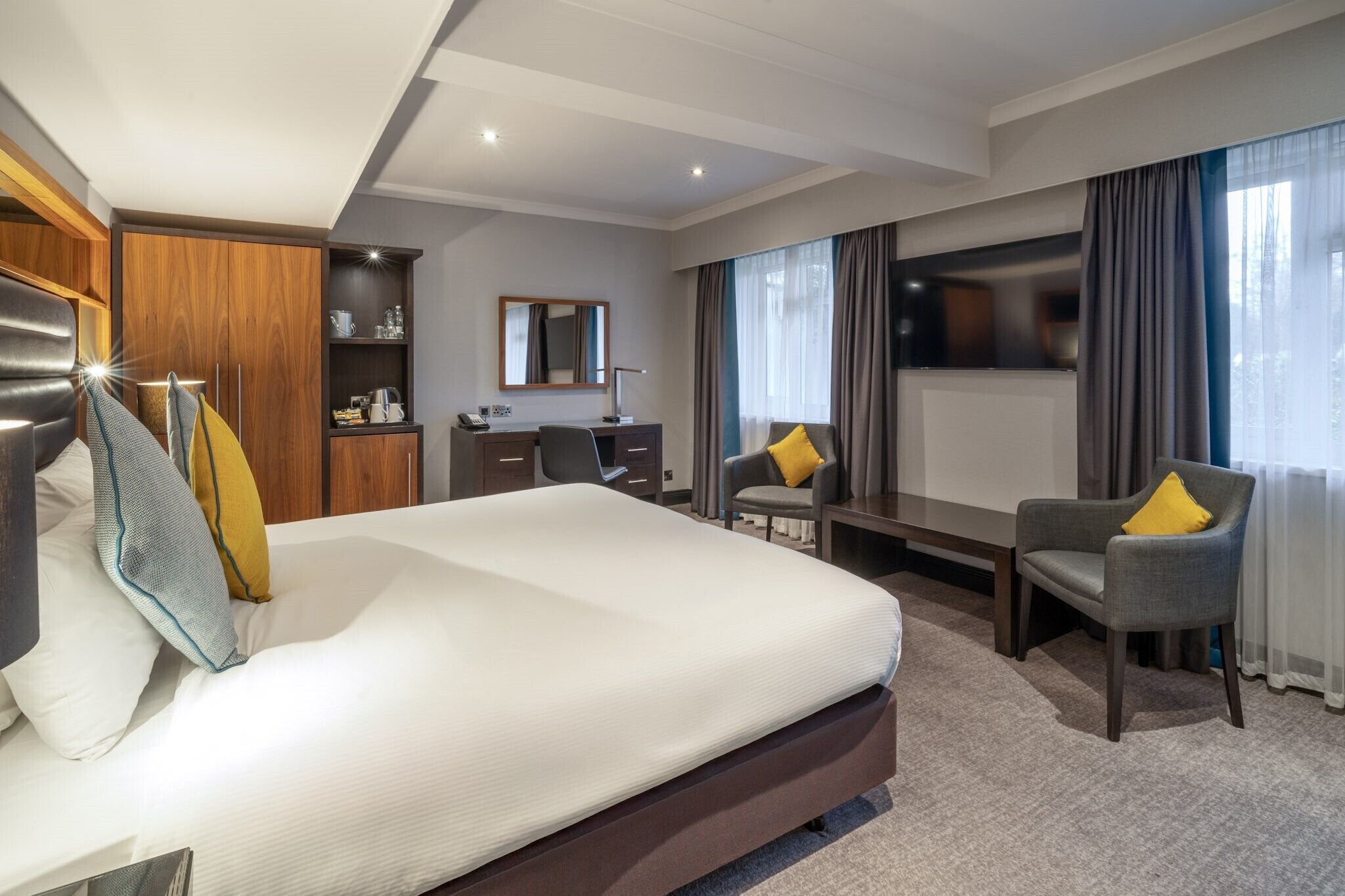A hotel room with a large bed, a desk, a chair, a wardrobe, and a TV.