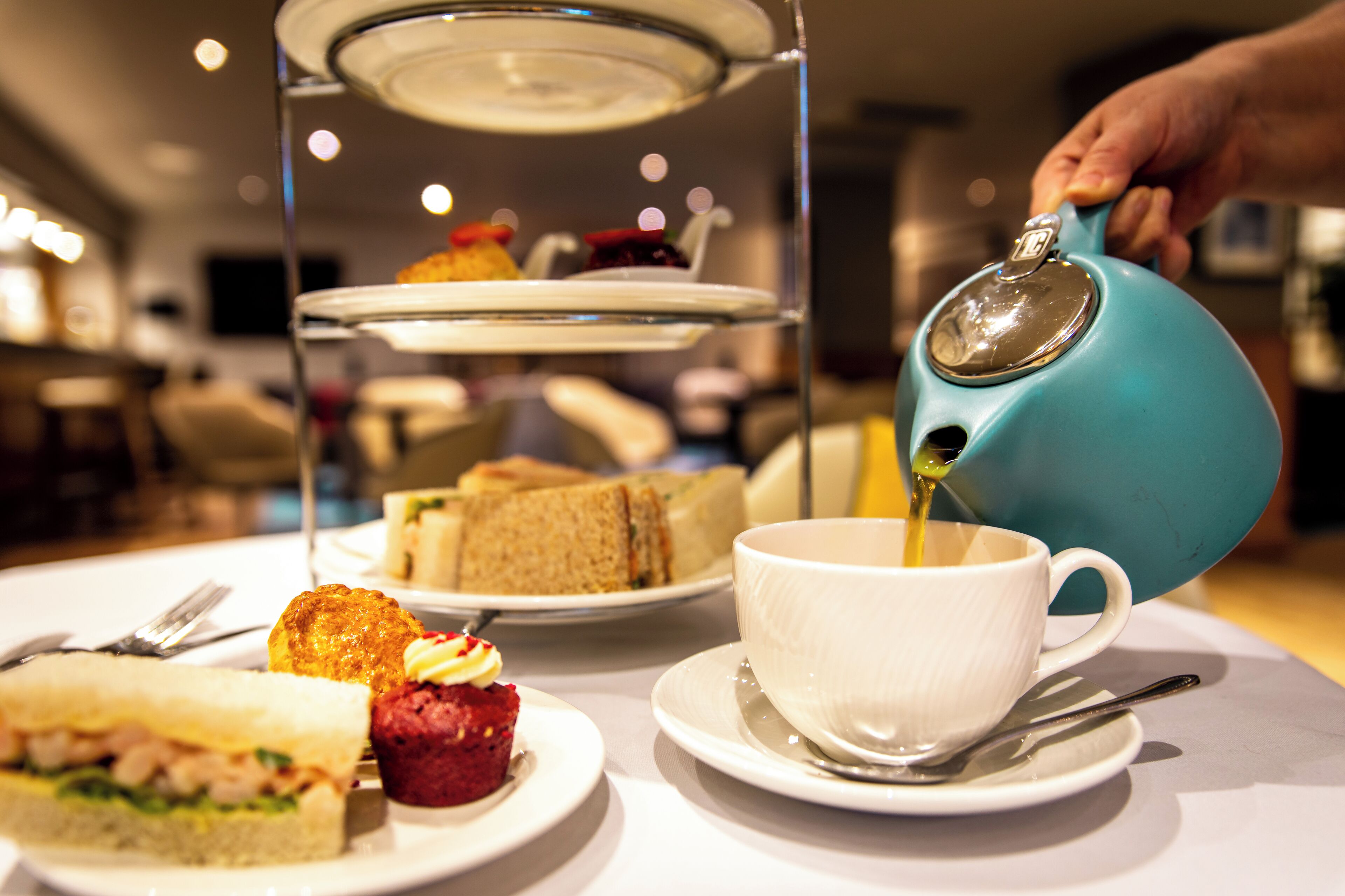 Un service à thé comprenant une théière bleue versant son contenu dans une tasse blanche, accompagné de divers sandwichs et d’un petit gâteau sur une table.