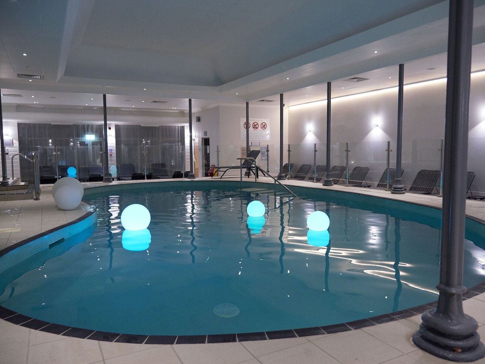 Piscina interior com água cristalina, rodeada por espreguiçadeiras e uma estrutura de vidro.