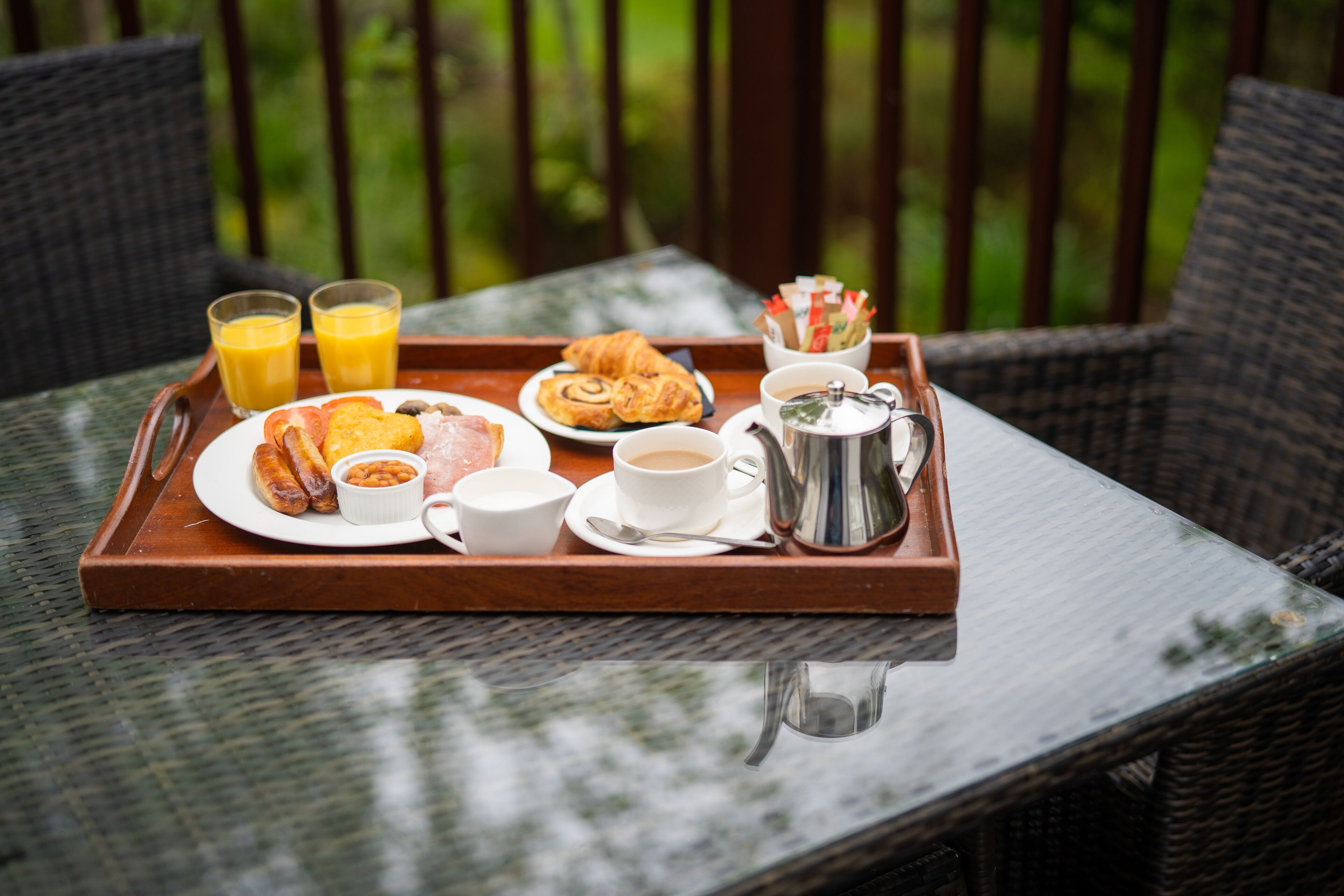 Bandeja de desayuno con croissants, salchichas, frijoles, café y jugo, sobre una mesa de mimbre con vista a la vegetación.