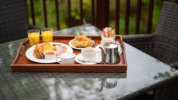 Bandeja de desayuno con croissants, salchichas, frijoles, café y jugo, sobre una mesa de mimbre con vista a la vegetación.