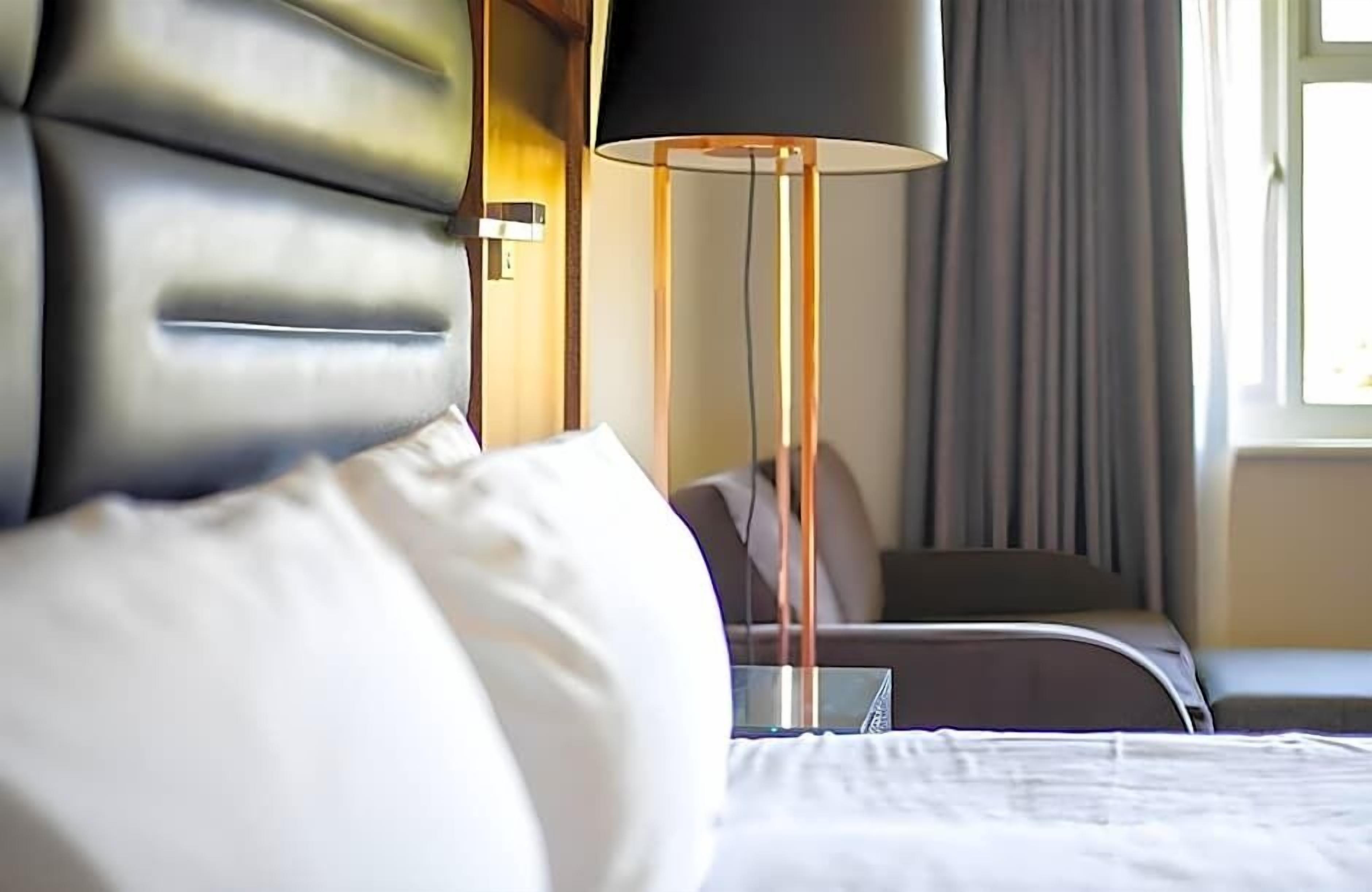 A hotel room with a bed, a bedside table, a lamp, and a window with curtains.