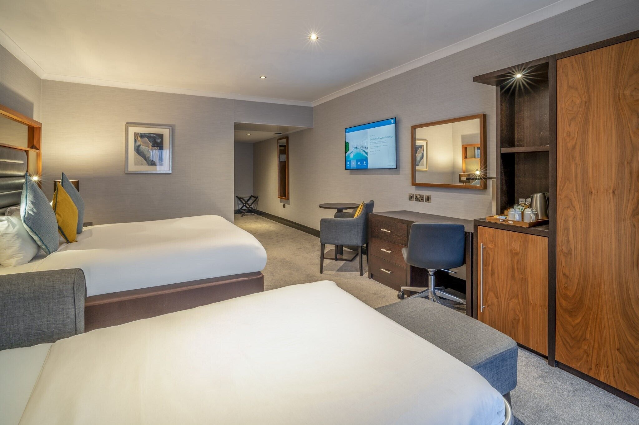 Habitación de hotel con dos camas, un escritorio, una silla, un armario y un televisor.
