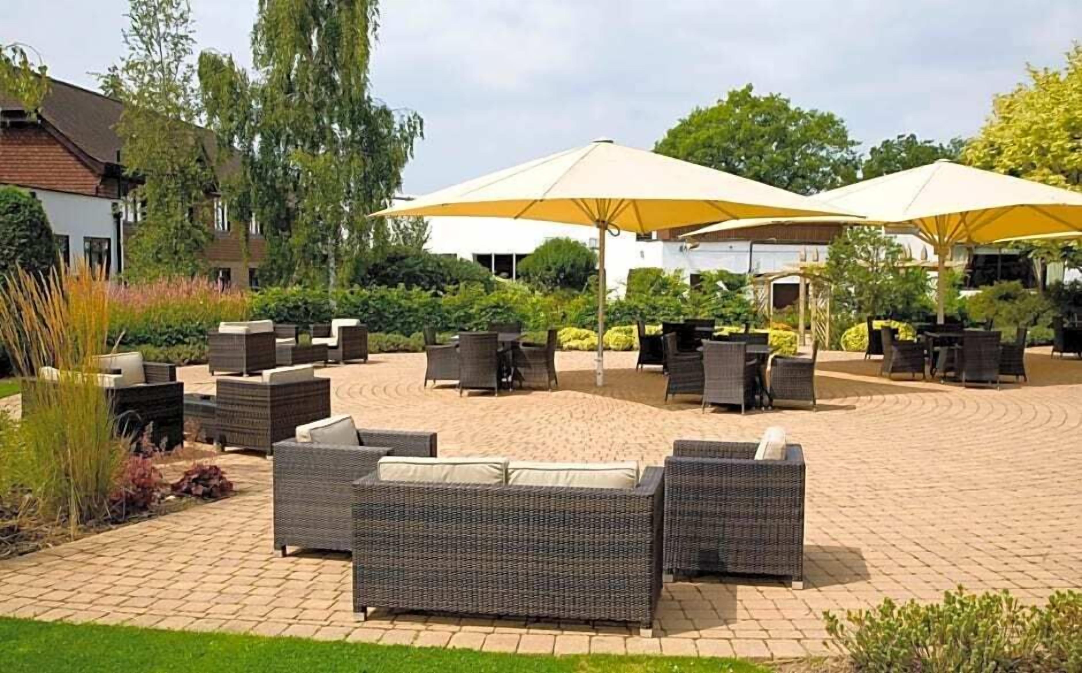 An outdoor seating area with wicker furniture and yellow umbrellas, surrounded by greenery and a paved walkway.