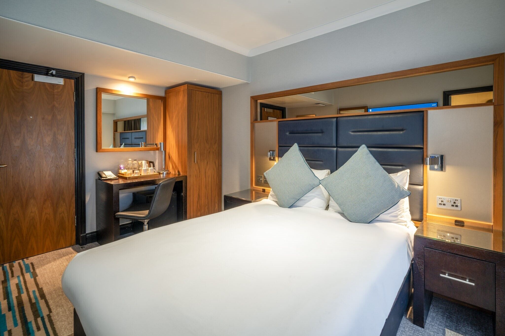 Quarto de hotel com uma cama grande, uma mesa com cadeira, um guarda-roupa e um espelho.