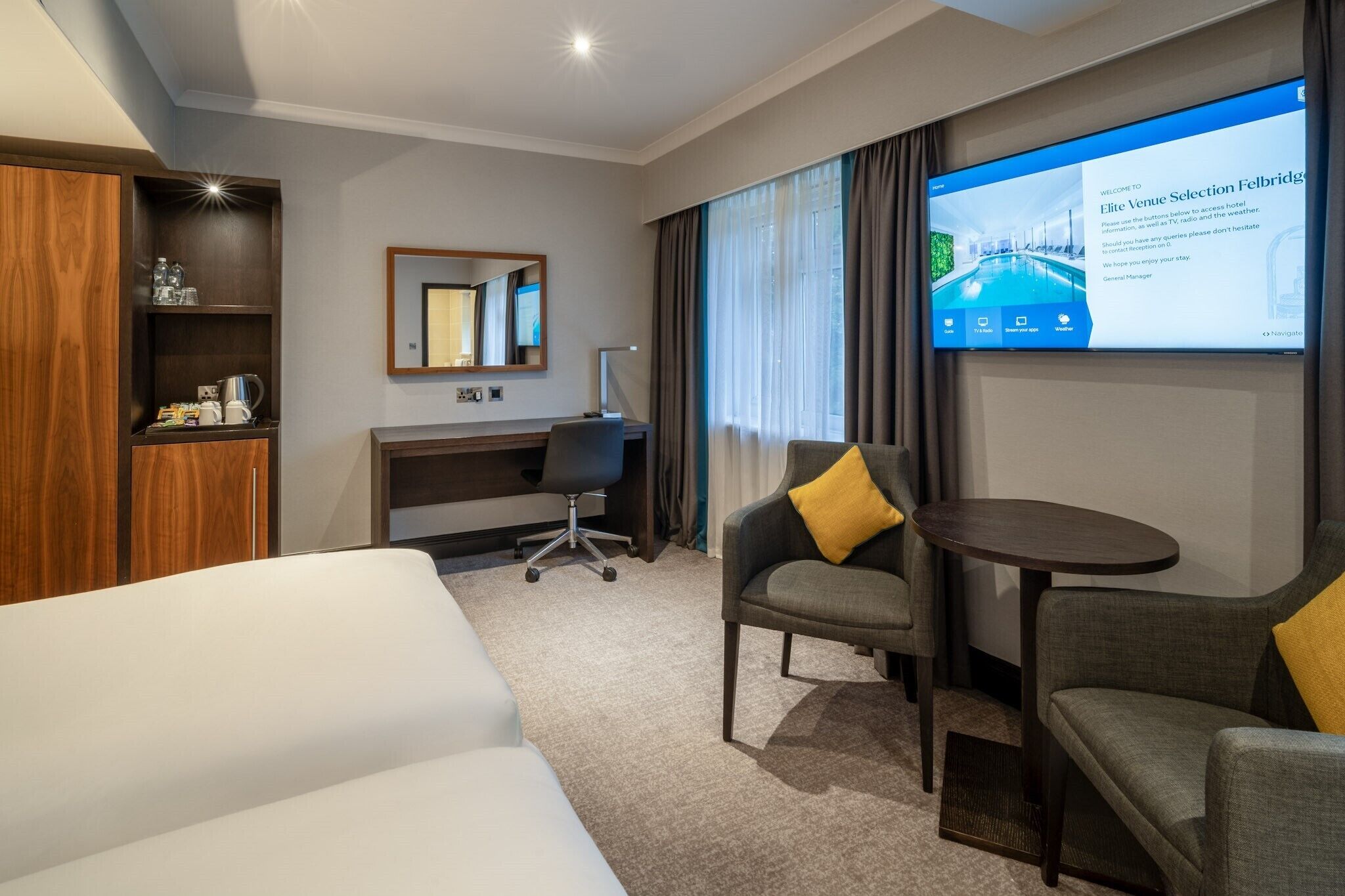 Ein Hotelzimmer mit einem Bett, einem Schreibtisch, einem Sessel, einem kleinen Tisch, einem Fernsehbildschirm und einem Holzkoffer.